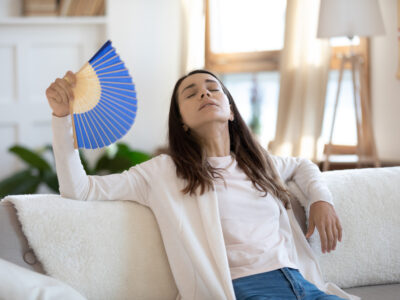 Frustrated unhappy young mixed race girl sitting on sofa, using paper fan, suffering from sunstroke or high temperature indoors, feeling unwell at summertime without air conditioning system at home.
