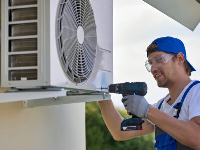 Smiling caucasian worker installs the outer metal case of the air conditioner with a screwdriver. Qualified male installer in uniform fixes air conditioner mount with tool